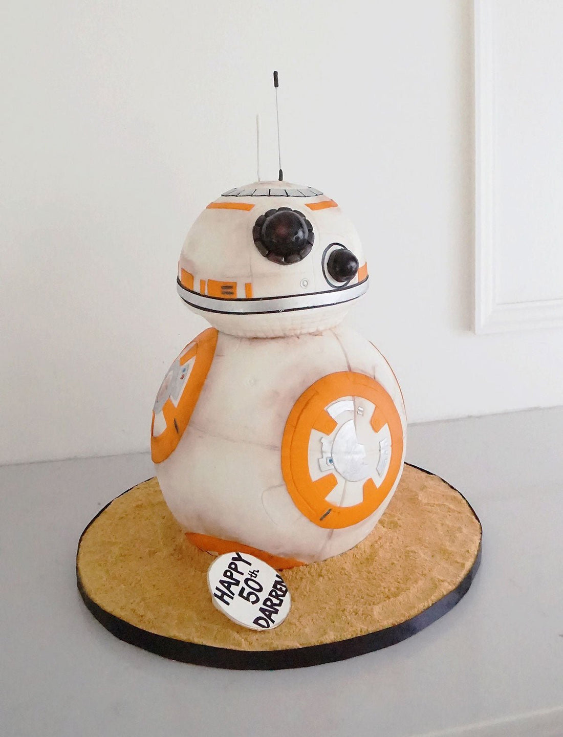 A cake shaped like BB-8, the droid from Star Wars, with white and orange details. It sits on a round sand-colored base with a small sign reading Happy 5th Birthday, Darcey.