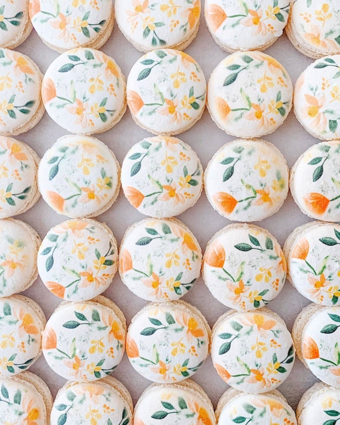 Neatly arranged on a light surface, Jenna Rae Cakes Printed Macarons feature white tops adorned with hand-painted orange flowers and green leaves.