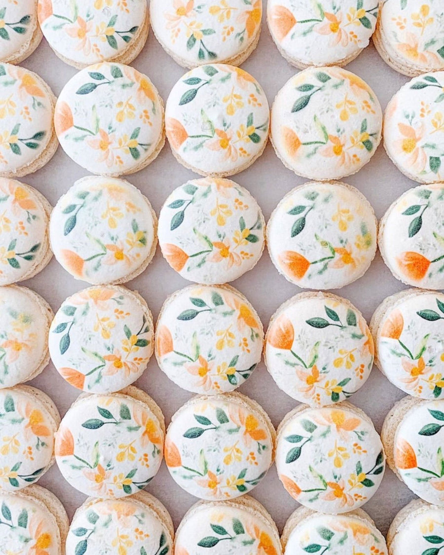 Neatly arranged on a light surface, Jenna Rae Cakes Printed Macarons feature white tops adorned with hand-painted orange flowers and green leaves.