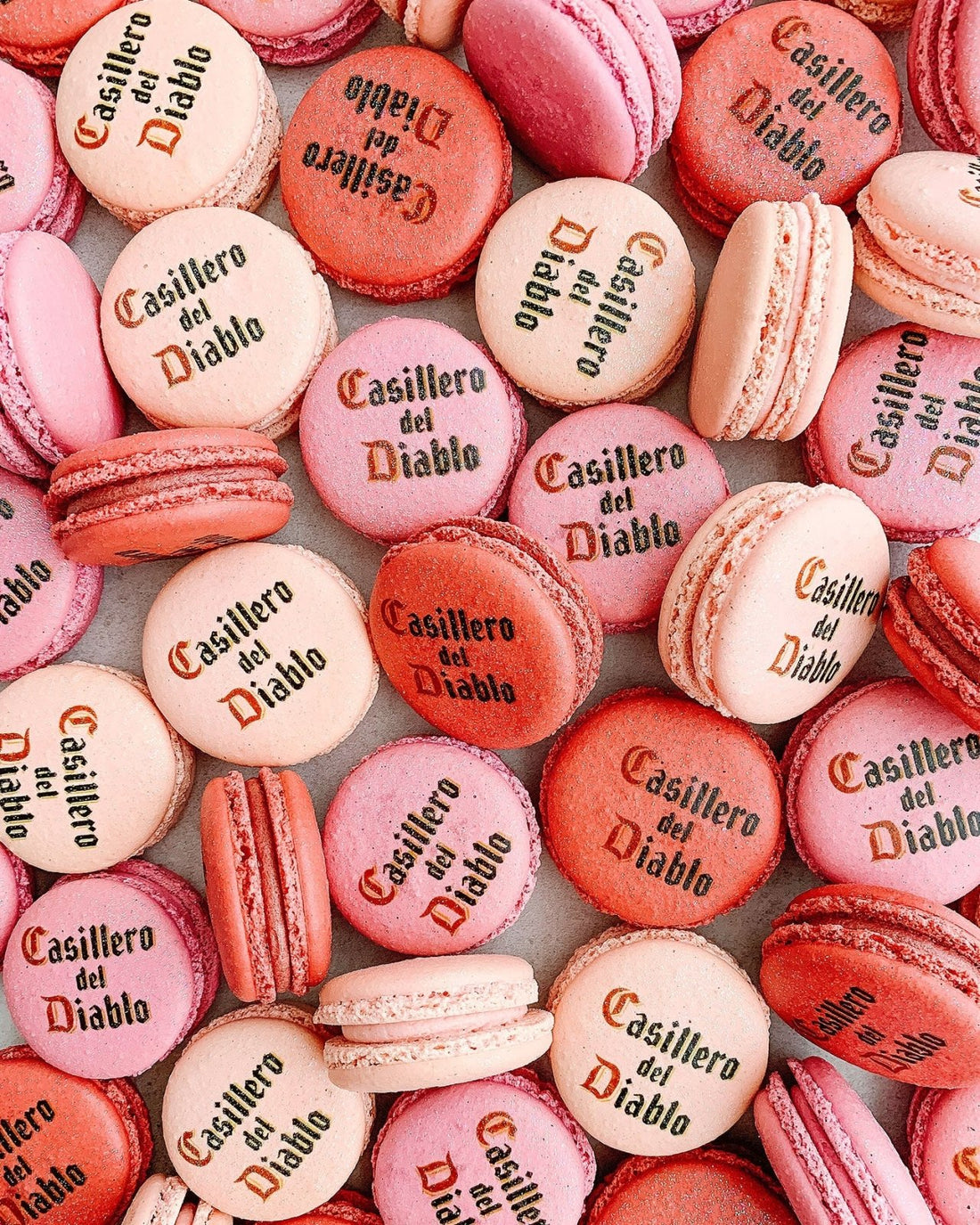 A pile of pink and red Printed Macarons by Jenna Rae Cakes, some featuring black lettering on top, are arranged close together to create a colorful and textured pattern.