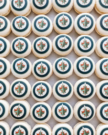 A grid of Printed Macarons by Jenna Rae Cakes, each topped with the Winnipeg Jets logo featuring a red maple leaf and jet in a blue and white circle.