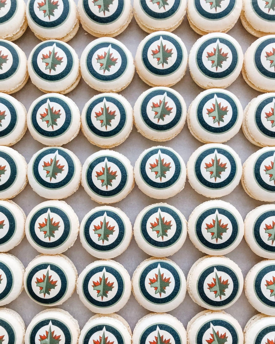 A grid of Printed Macarons by Jenna Rae Cakes, each topped with the Winnipeg Jets logo featuring a red maple leaf and jet in a blue and white circle.