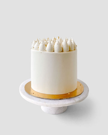 The Red Velvet Cake by Jenna Rae Cakes features white frosting with piped dollops and gold flakes, presented on a white marble cake stand with a gold base against a light gray background.