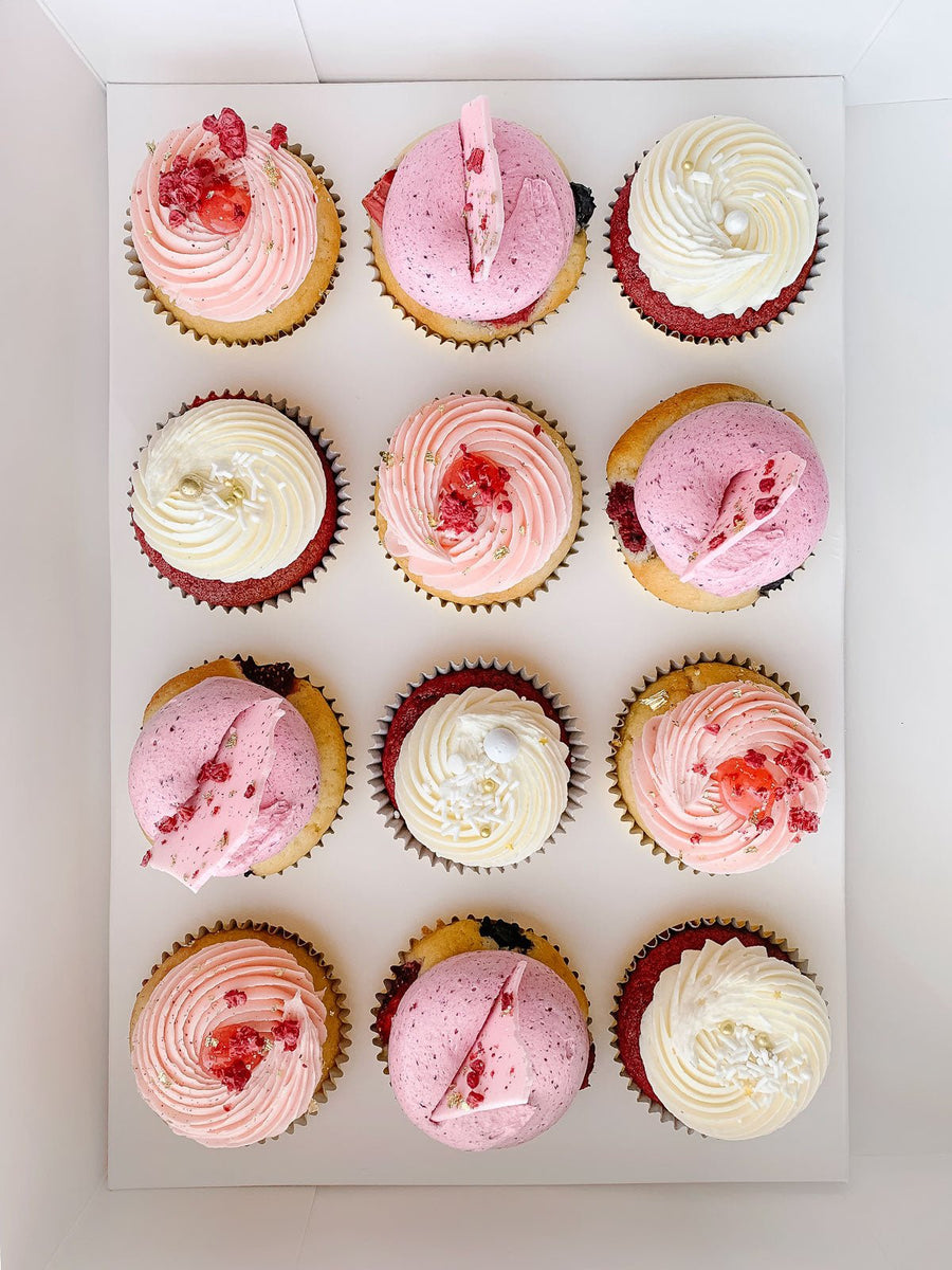 Enjoy a box of twelve Regular Cupcakes from Jenna Rae Cakes, each topped with pink, white, and purple swirled frosting, sprinkles, and finished with dried fruit or chocolate pieces.