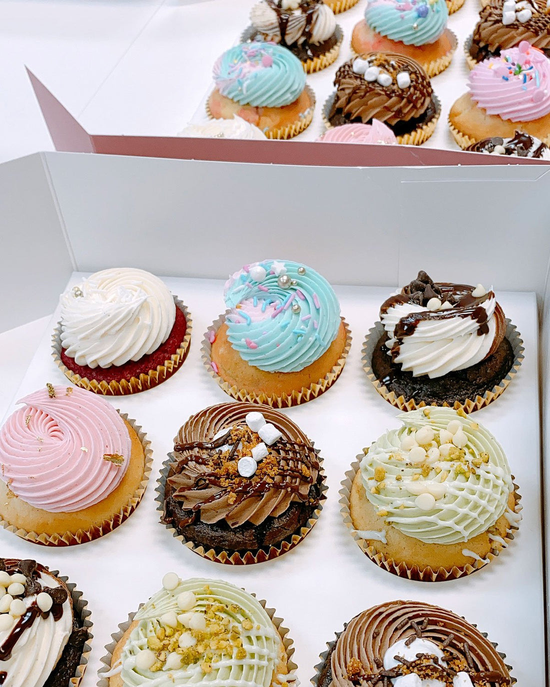 An open box and tray of Jenna Rae Cakes Regular Cupcakes, each topped with colorful swirled frosting, sprinkles, and decorative toppings, arranged neatly in rows.