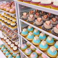 Jenna Rae Cakes Regular Cupcakes, featuring colorful icing swirls in white, pink, blue, and chocolate and some topped with strawberries, are neatly arranged on metal racks in a bakery display.