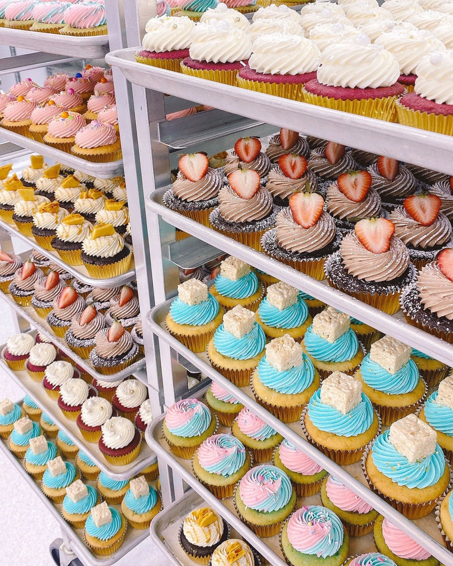 Jenna Rae Cakes Regular Cupcakes, featuring colorful icing swirls in white, pink, blue, and chocolate and some topped with strawberries, are neatly arranged on metal racks in a bakery display.