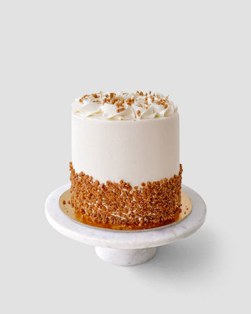 The Salted Caramel Crunch Cake by Jenna Rae Cakes is a round, white frosted cake with whipped cream swirls and crushed nuts around the base, displayed on a marble stand against a plain light background.