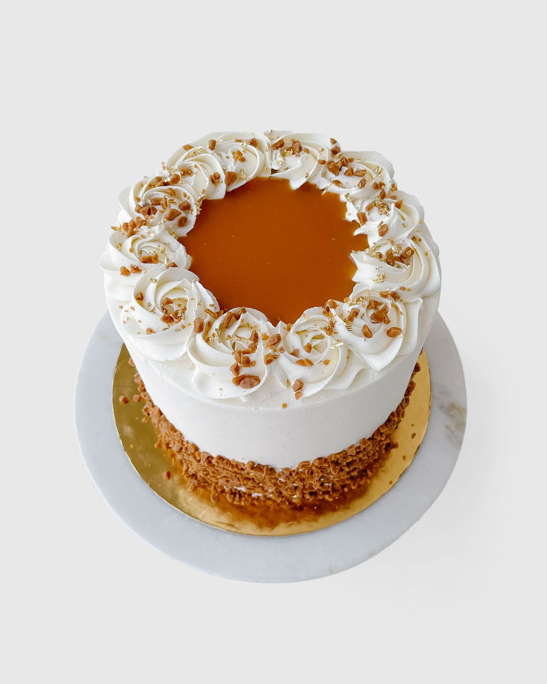 The Salted Caramel Crunch Cake by Jenna Rae Cakes features white frosting, caramel sauce, decorative swirls, and crushed nuts around the edges. It’s presented on a gold board atop a white marble surface.