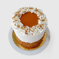 The Salted Caramel Crunch Cake by Jenna Rae Cakes features white frosting, caramel sauce, decorative swirls, and crushed nuts around the edges. It’s presented on a gold board atop a white marble surface.