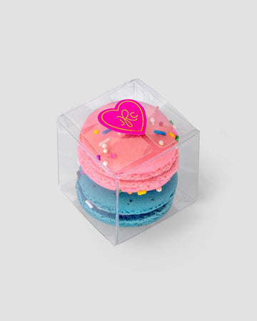 Jenna Rae Cakes’ Signature 2-Macaron Box features a clear plastic container with pink and blue macarons stacked inside, topped with colorful sprinkles and a pink heart sticker, set against a light gray background.
