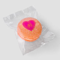 The Jenna Rae Cakes Signature Single Macaron Bag features one orange macaron in a clear wrapper, sealed with a pink heart sticker with yellow letters, displayed on a plain light gray background.