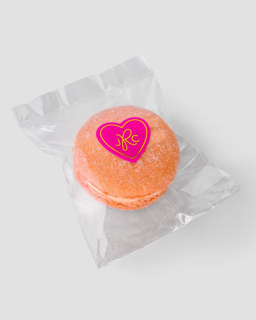 The Jenna Rae Cakes Signature Single Macaron Bag features one orange macaron in a clear wrapper, sealed with a pink heart sticker with yellow letters, displayed on a plain light gray background.