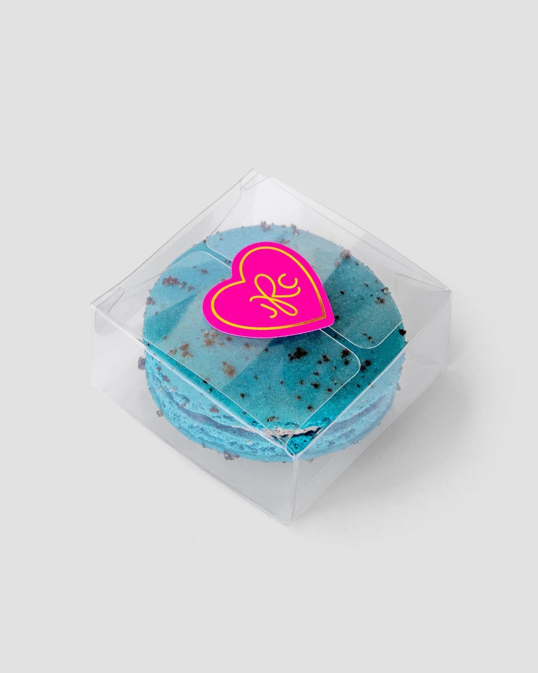 The Signature Single Macaron Box by Jenna Rae Cakes showcases a blue macaron with chocolate specks inside a clear plastic box, sealed with a pink heart-shaped sticker featuring a yellow design, set against a plain light gray background.