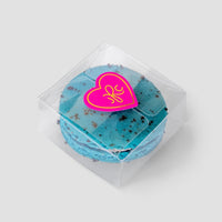 The Signature Single Macaron Box by Jenna Rae Cakes showcases a blue macaron with chocolate specks inside a clear plastic box, sealed with a pink heart-shaped sticker featuring a yellow design, set against a plain light gray background.
