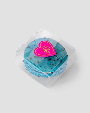 The Signature Single Macaron Box by Jenna Rae Cakes showcases a blue macaron with chocolate specks inside a clear plastic box, sealed with a pink heart-shaped sticker featuring a yellow design, set against a plain light gray background.