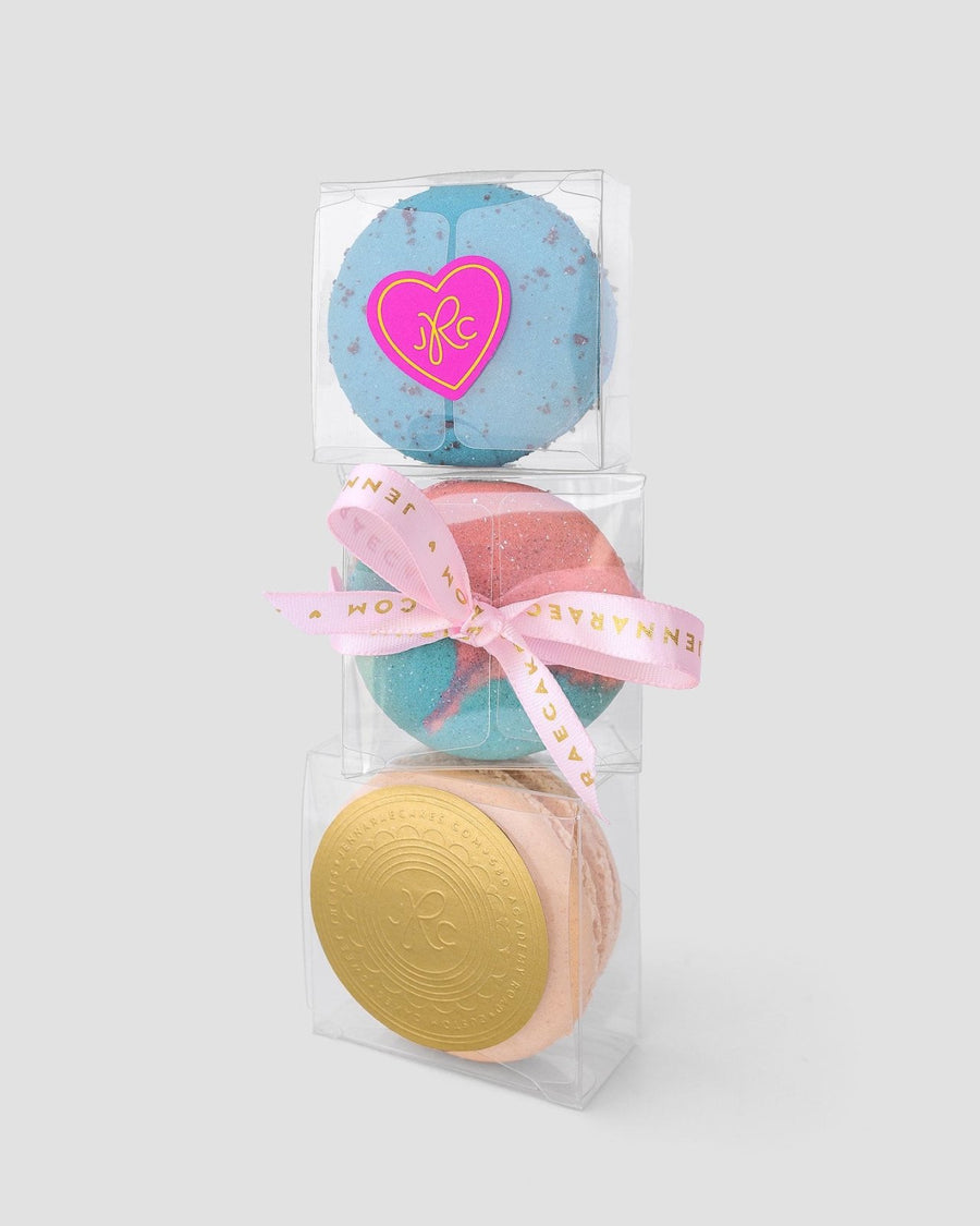 Three stacked Signature Single Macaron Boxes by Jenna Rae Cakes: the top is blue with a heart-shaped sticker, the middle is pink and blue with a pink ribbon, and the bottom is peach with a round gold embossed label.