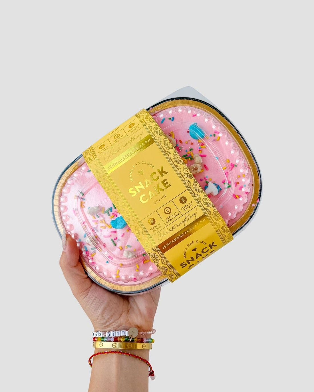 A hand with colorful beaded bracelets holds a Jenna Rae Cakes Snack Cake, featuring pink frosting, colorful sprinkles, and wrapped in a yellow label that reads Snack Cakes.