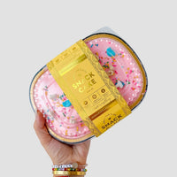 A hand with colorful beaded bracelets holds a Jenna Rae Cakes Snack Cake, featuring pink frosting, colorful sprinkles, and wrapped in a yellow label that reads Snack Cakes.