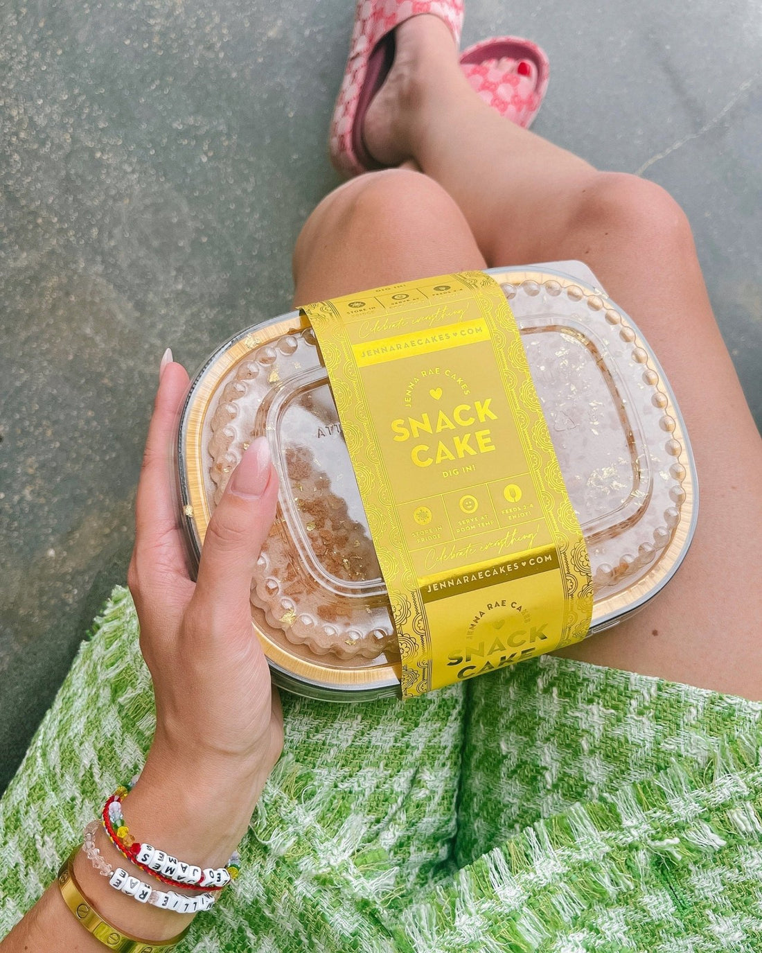 A person in a green textured skirt holds a clear plastic Snack Cakes container by Jenna Rae Cakes with a yellow wrapper. They wear colorful beaded bracelets, pink checkered shoes, and sit cross-legged.