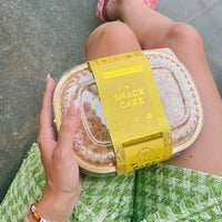 A person in a green textured skirt holds a clear plastic Snack Cakes container by Jenna Rae Cakes with a yellow wrapper. They wear colorful beaded bracelets, pink checkered shoes, and sit cross-legged.