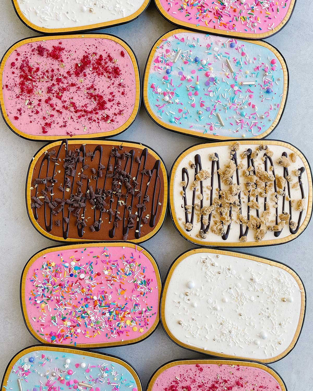 Boxes of Jenna Rae Cakes Snack Cakes are arranged in a grid, showcasing flavors like pink with sprinkles, blue with confetti, chocolate drizzle, cookie dough, white with pearls, and others topped with treats.