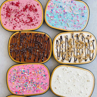 Boxes of Jenna Rae Cakes Snack Cakes are arranged in a grid, showcasing flavors like pink with sprinkles, blue with confetti, chocolate drizzle, cookie dough, white with pearls, and others topped with treats.