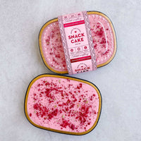 Two small oval containers of Jenna Rae Cakes Snack Cakes, one labeled and the other open to reveal pink cake with frosting and red sprinkles, displayed on a light gray surface.