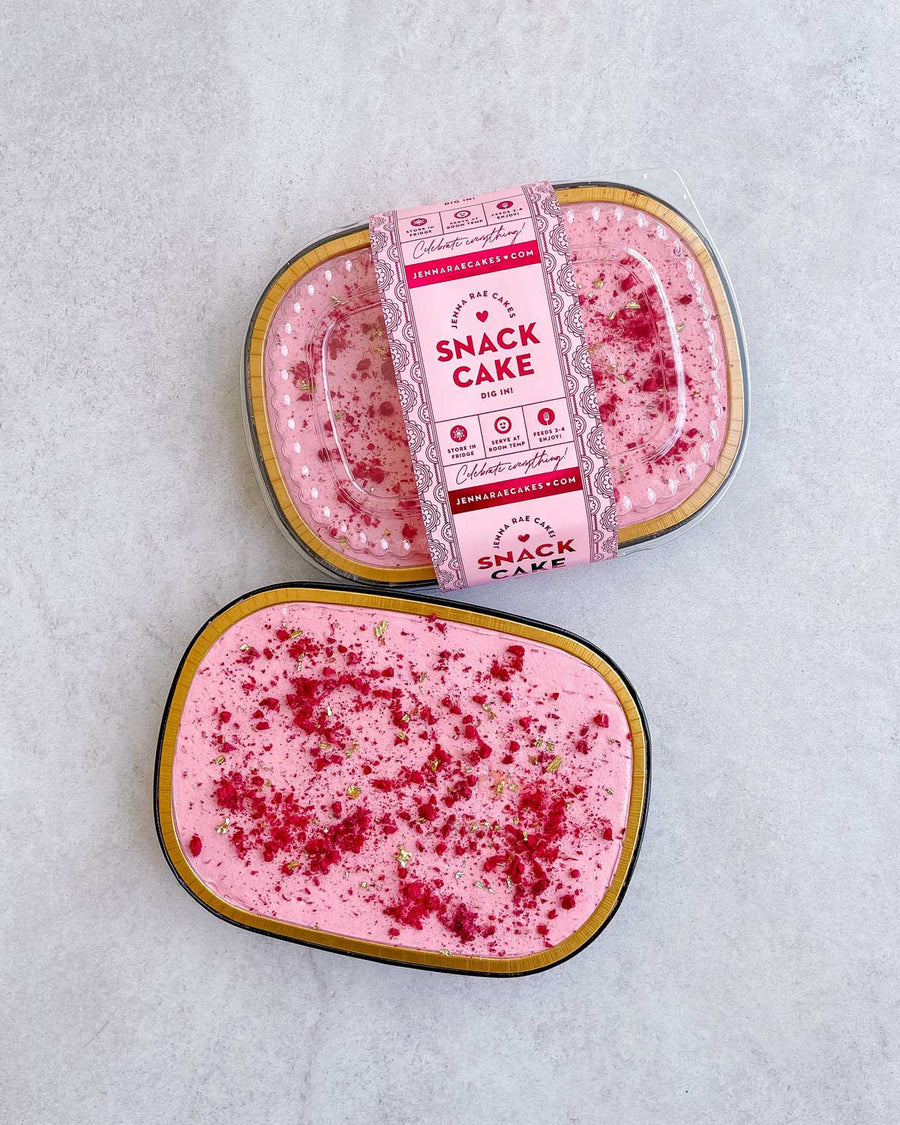 Two small oval containers of Jenna Rae Cakes Snack Cakes, one labeled and the other open to reveal pink cake with frosting and red sprinkles, displayed on a light gray surface.