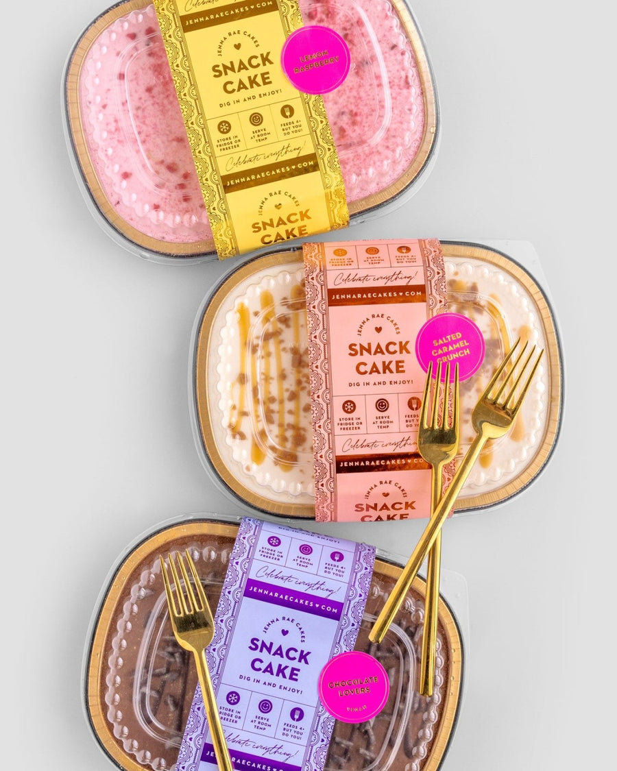 Three packaged Snack Cakes by Jenna Rae Cakes—pink, caramel, and chocolate—each with decorative labels and a gold fork on top, arranged on a light grey background.