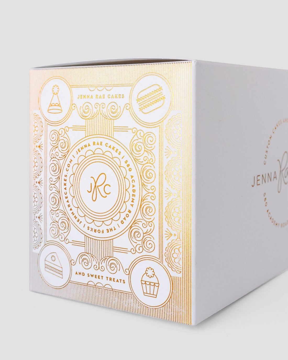 The Sweet Treat Gift Box by Jenna Rae Cakes is a white bakery box with gold embossed cake, cupcake, and macaron icons, featuring the brand’s logo and elegant decorative patterns.