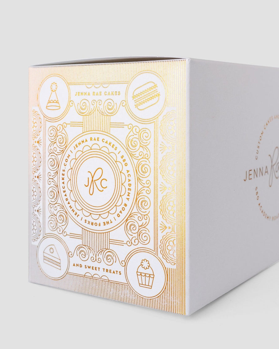 The Sweet Treat Gift Box by Jenna Rae Cakes is a white bakery box with gold embossed cake, cupcake, and macaron icons, featuring the brand’s logo and elegant decorative patterns.