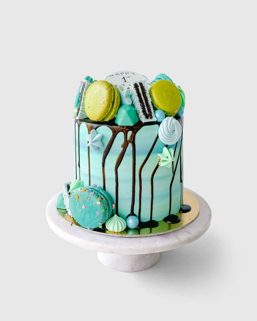 The Full Works Cake by Jenna Rae Cakes is a tall, blue-green ombre cake with chocolate drip, topped with assorted macarons, meringues, and cookies, displayed on a white cake stand against a plain background.