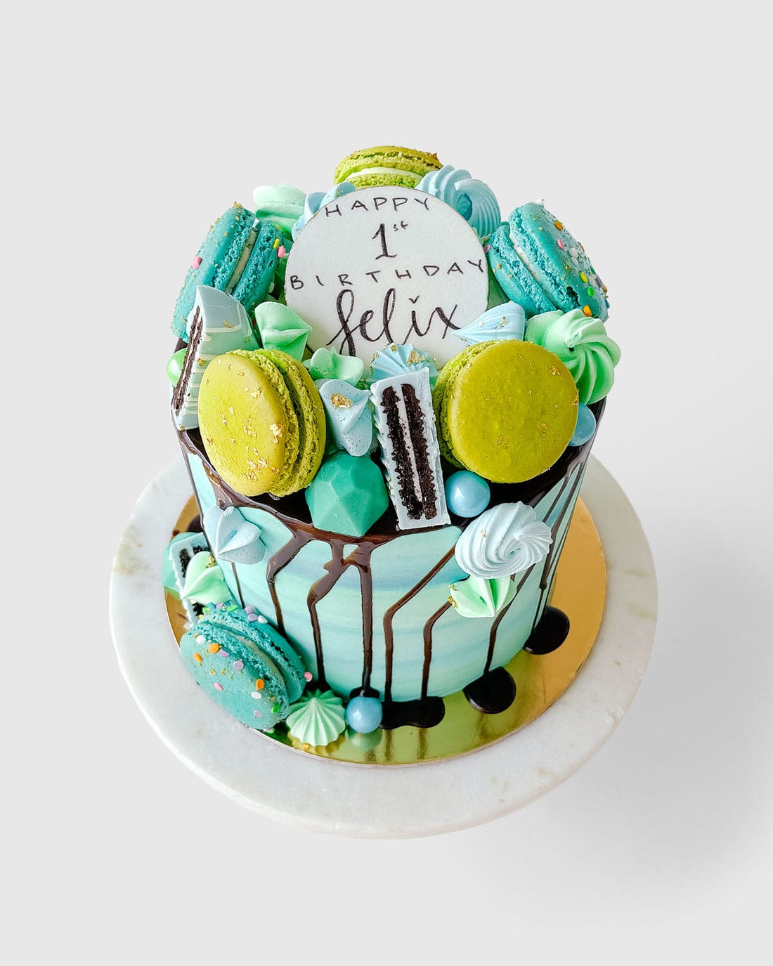 The Full Works Cake by Jenna Rae Cakes is a tall blue and green birthday cake topped with macarons, meringues, chocolates, and a large cookie reading “Happy 1st Birthday Felix,” finished with chocolate drips on a marble plate.