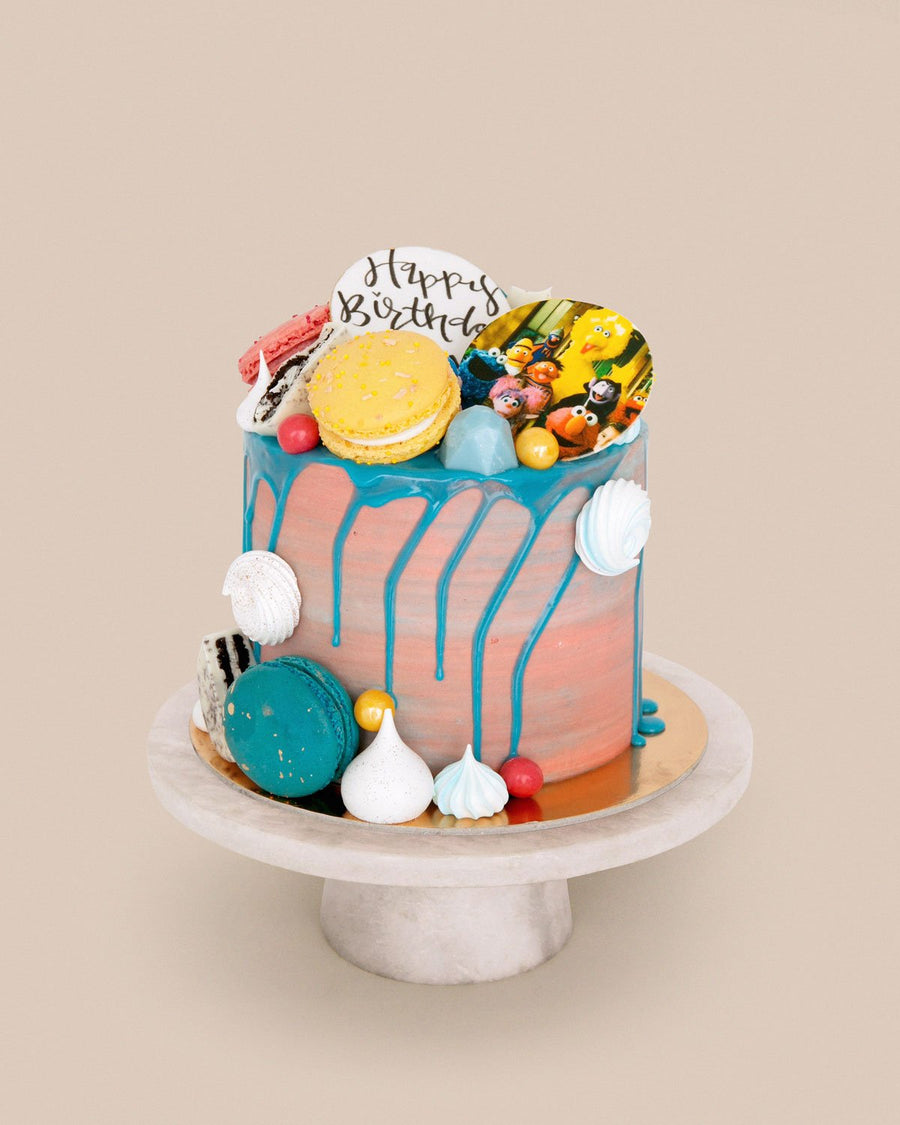 The Kids Works Cake by Jenna Rae Cakes features blue drip icing, macarons, meringues, candy balls, and a Happy Birthday sign, displayed on a white cake stand against a beige background.