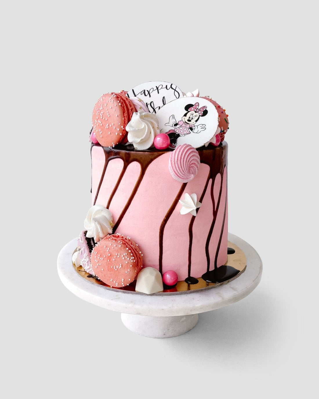 The Kids Works Cake by Jenna Rae Cakes is a pink cake with chocolate drip, topped with pink macarons, meringues, candy, and a white topper featuring a cartoon mouse and “Happy B,” displayed on a white stand against a plain backdrop.