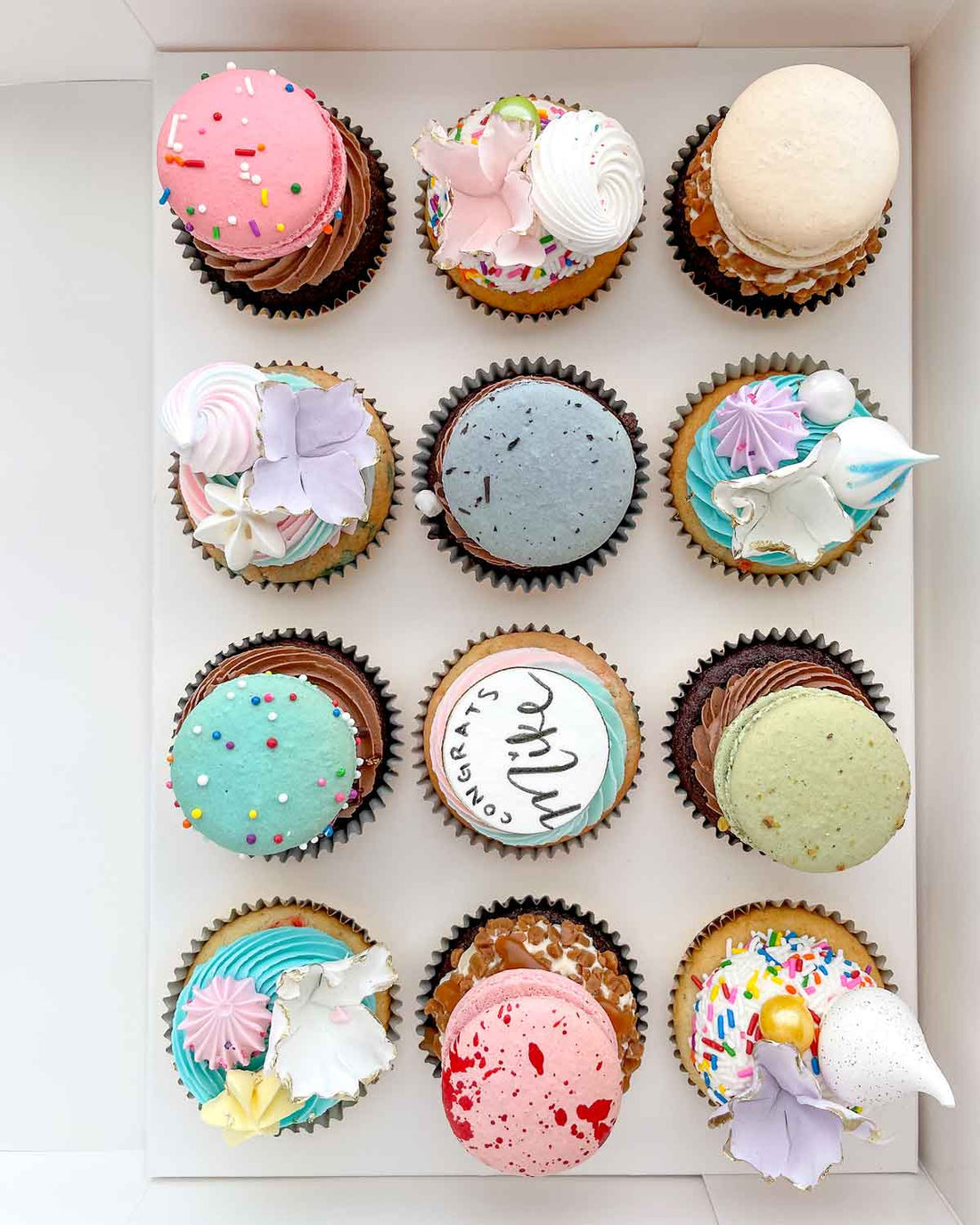 The Works Cupcakes by Jenna Rae Cakes features 12 uniquely decorated cupcakes with colorful frosting, sprinkles, macarons, flowers, and edible pearls, neatly arranged in three rows of four.
