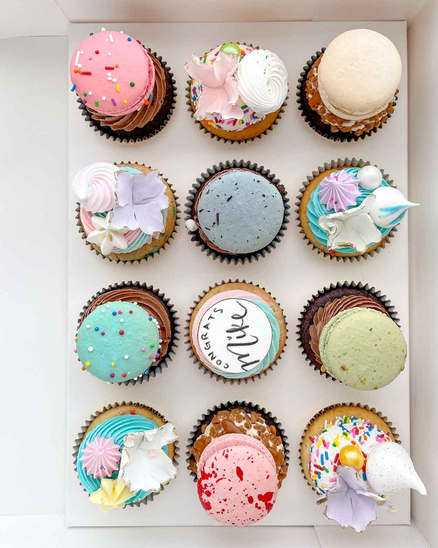The Works Cupcakes by Jenna Rae Cakes features 12 uniquely decorated cupcakes with colorful frosting, sprinkles, macarons, flowers, and edible pearls, neatly arranged in three rows of four.