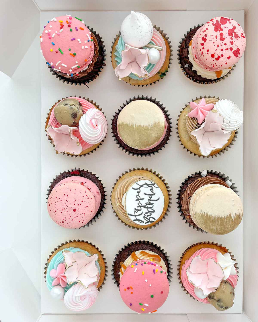 The Works Cupcakes by Jenna Rae Cakes: a box of 12 assorted cupcakes topped with macarons, gold accents, sprinkles, meringue, and pink flower-shaped icing. One cupcake features Happy Birthday in black script.