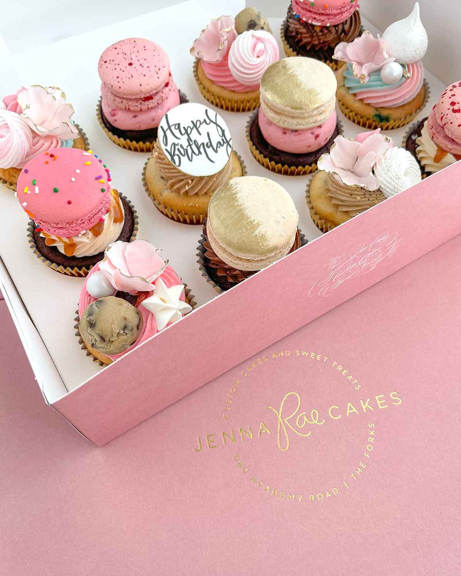 The Works Cupcakes by Jenna Rae Cakes come in a pink box with a gold logo and include assorted cupcakes topped with pink and gold frosting, macarons, cookie dough, meringues, and a “Happy Birthday” round topper.