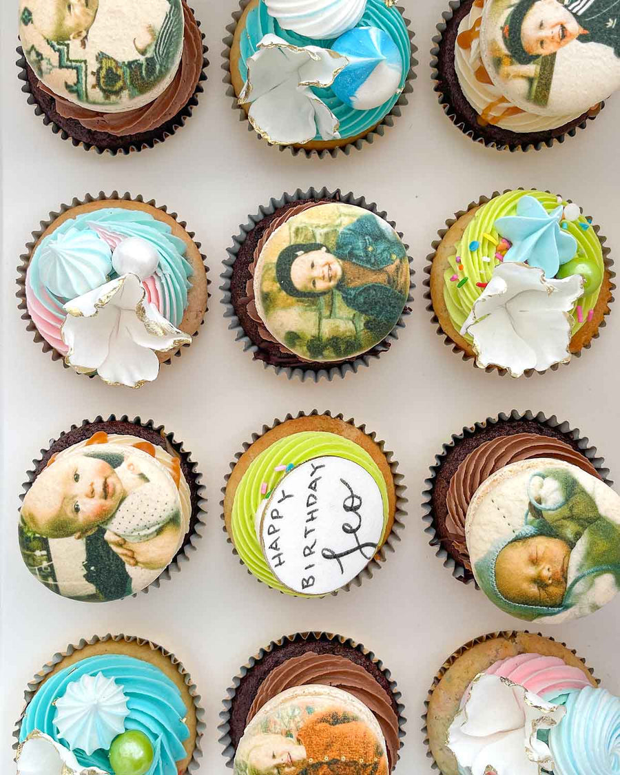 The Works Cupcakes by Jenna Rae Cakes: a box of colorful cupcakes, some with pastel swirls and white chocolate shards, others featuring edible baby and child images, plus one with Happy Birthday Joe on top.