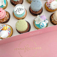 An open pink Jenna Rae Cakes box displays The Works Cupcakes, each decorated with colorful macarons, meringues, sprinkles, and a cookie that reads “Congrats Mike.”.