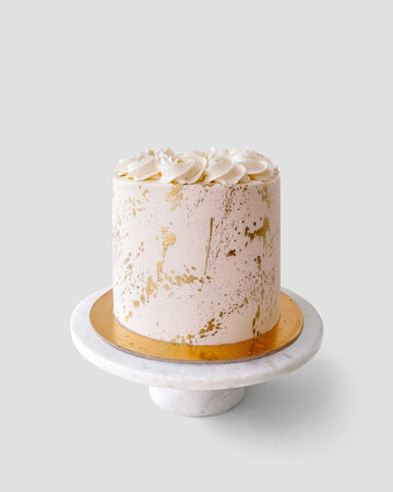 The Vanilla Cake by Jenna Rae Cakes features white frosting with gold splatters and cream swirls, presented on a gold board and white marble stand against a plain light background.