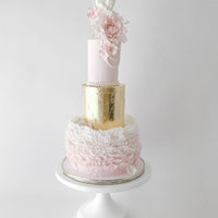 A three-tiered cake with pink, gold, and white layers; decorated with ruffled icing, gold leaf, pearls, and delicate pink and white sugar flowers, displayed on a white cake stand against a plain background.