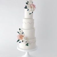 A four-tiered white wedding cake decorated with delicate pastel flowers and greenery, featuring subtle gold accents and pearl-like borders, displayed on a white stand against a plain background.