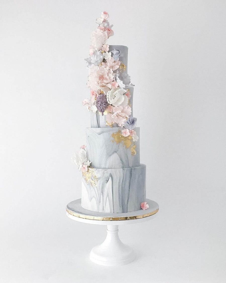 A four-tiered marble-patterned cake with gold accents, decorated with pastel pink, white, and purple sugar flowers cascading down one side, displayed on a white cake stand with a gold rim.