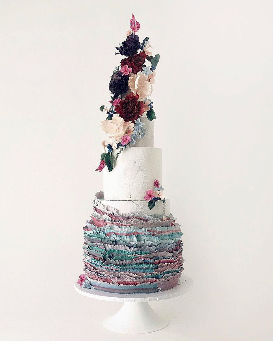 A three-tiered cake with textured, ruffled layers in pastel colors and decorated with cascading, realistic flowers in shades of pink, purple, and white, displayed on a white cake stand against a plain background.