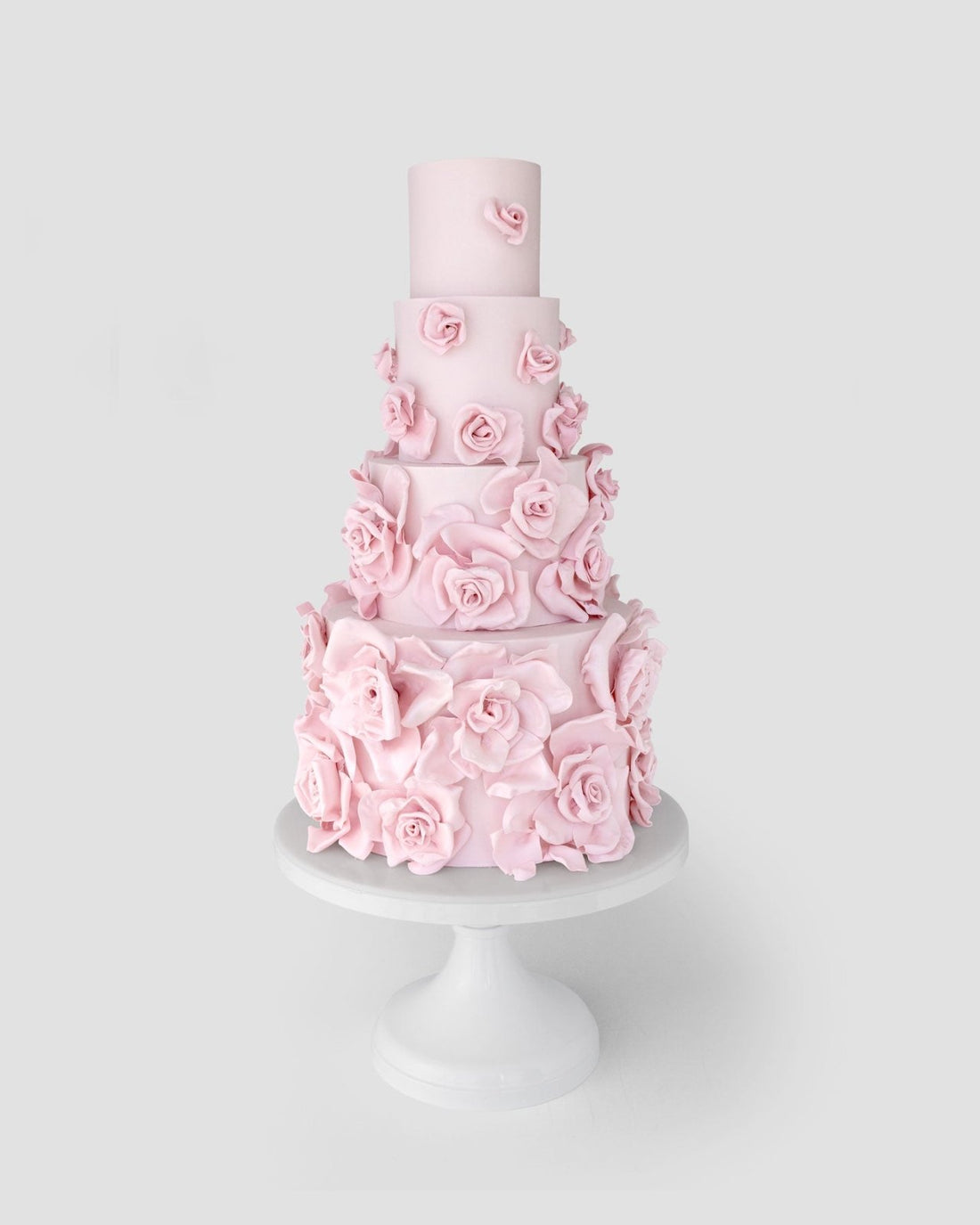 A tall, four-tiered cake decorated with numerous pastel pink fondant roses, displayed on a white pedestal cake stand against a light gray background.