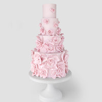 A tall, four-tiered cake decorated with numerous pastel pink fondant roses, displayed on a white pedestal cake stand against a light gray background.