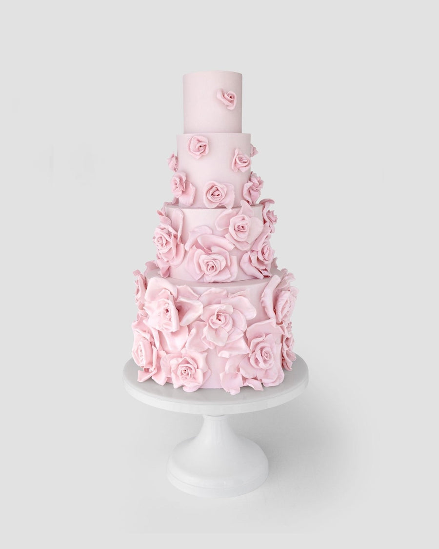 A tall, four-tiered cake decorated with numerous pastel pink fondant roses, displayed on a white pedestal cake stand against a light gray background.